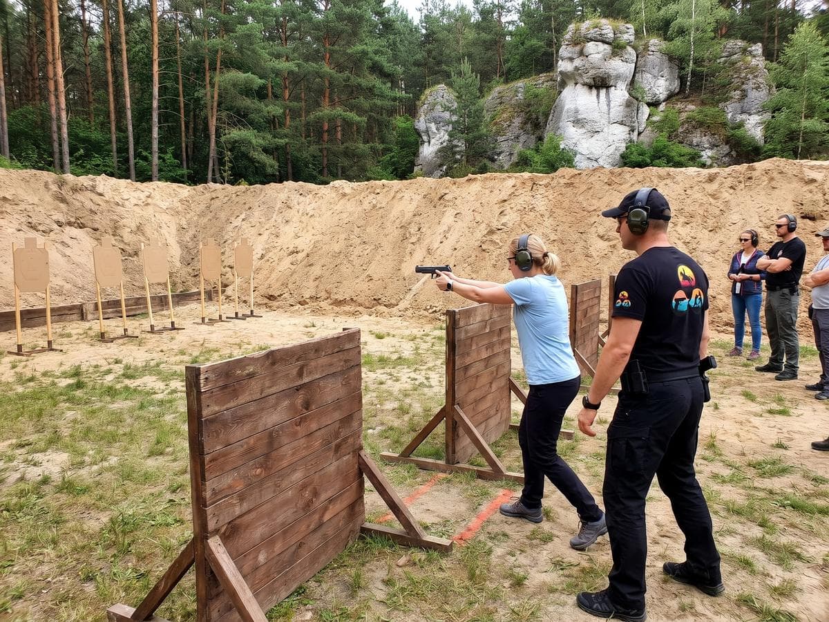 Uczestniczka strzela z pistoletu między drewnianymi barykadami na strzelnicy, a tuż za nią czuwa instruktor Horseback.